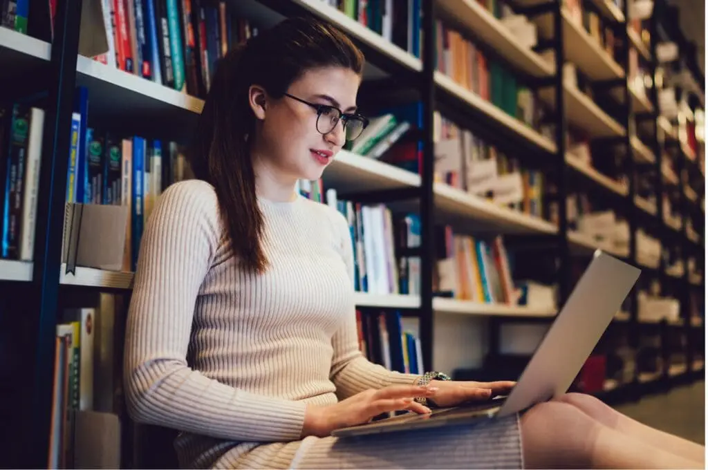A young college student accesses student support services on her laptop while sitting on the library floor.