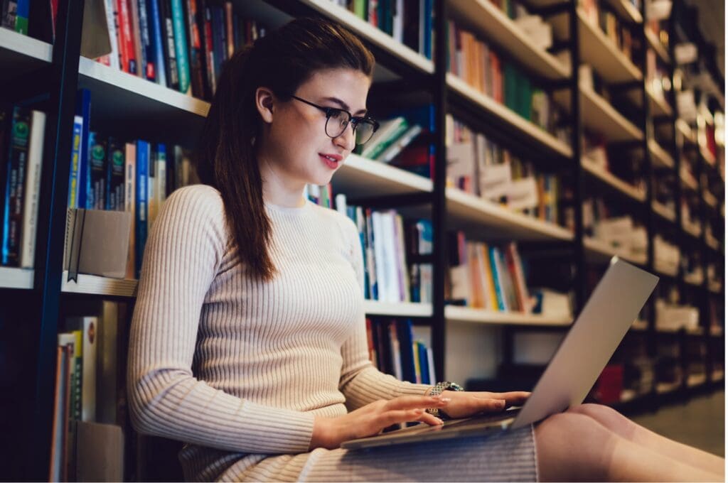 A young college student accesses student support services on her laptop while sitting on the library floor.