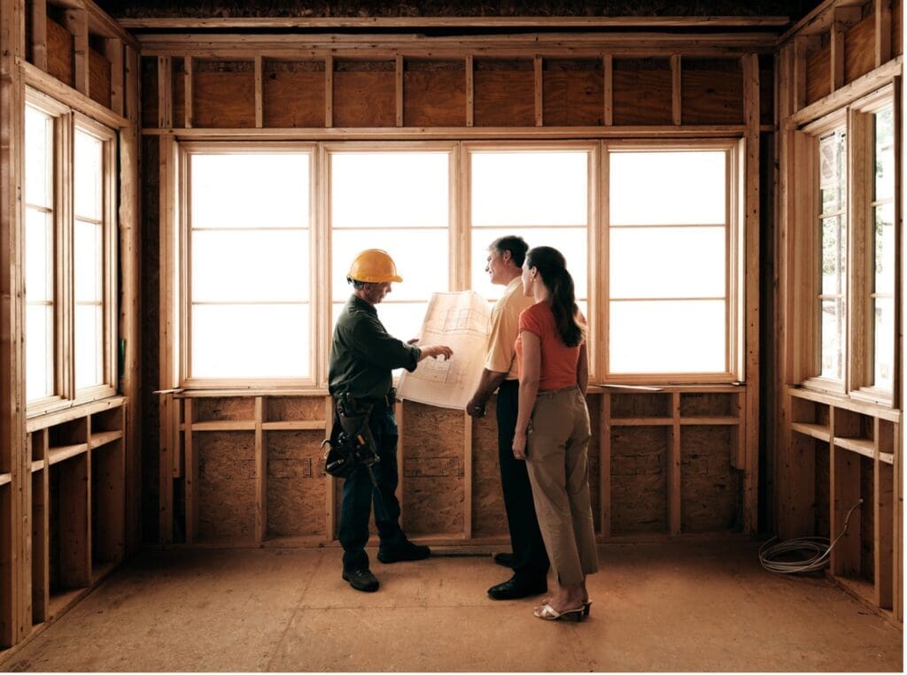 A residential builder stands in a home under construction consulting with homebuyers about affordability.
