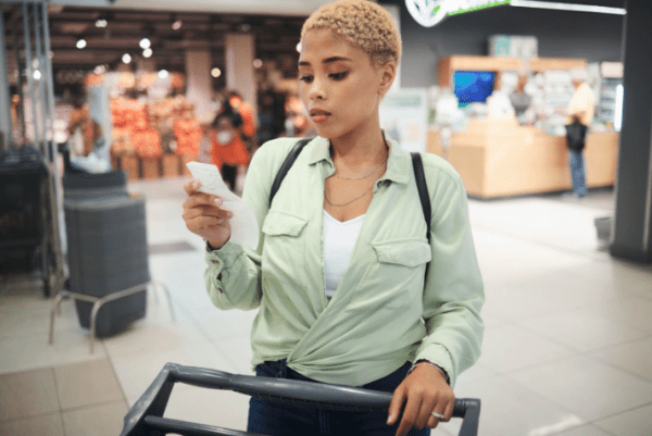 Woman in green shirt walking through a store pushing a shopping cart looking at her cell phone. 