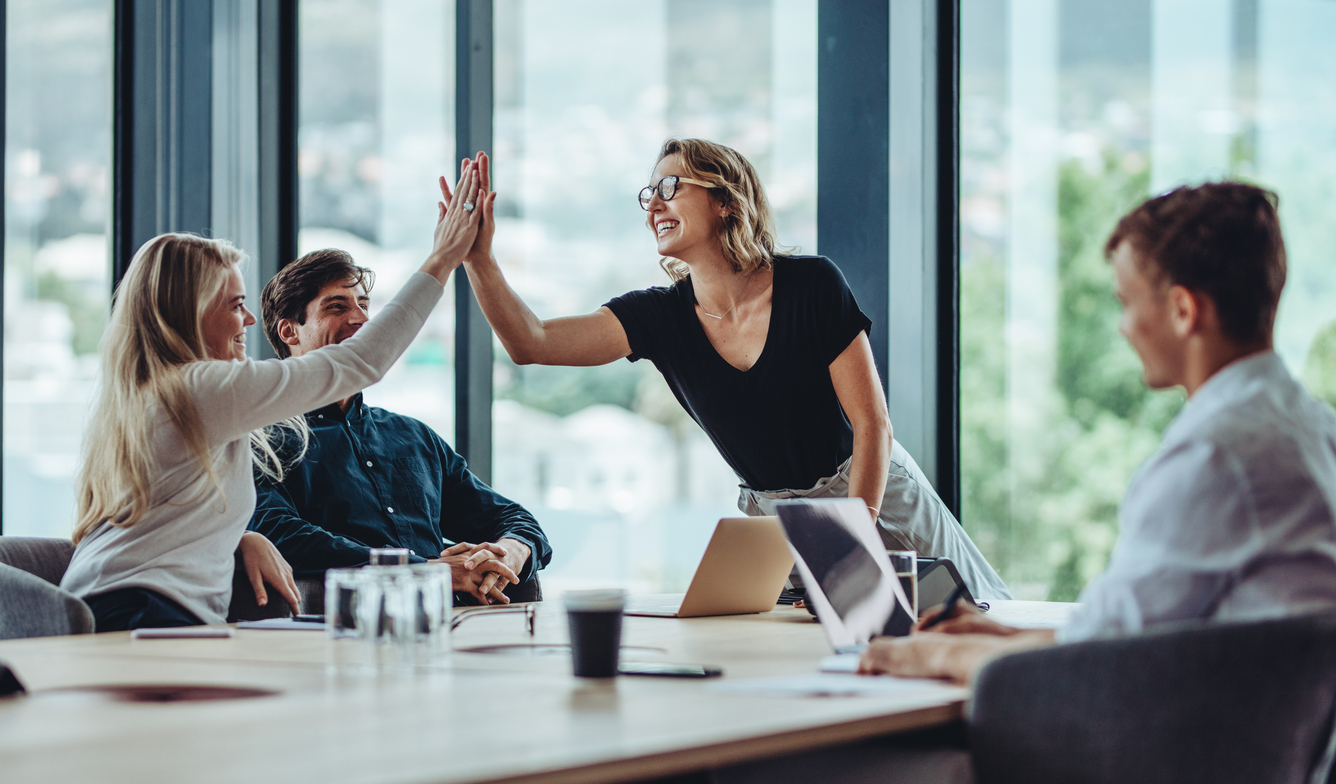 A happy sales team sits around a table, giving high fives over increased conversion rates that resulted from monitoring lead qualification metrics. 