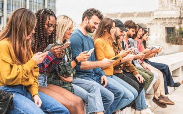 A group of people sit on a wall checking messages on their phones, illustrating the rise in mass texting.