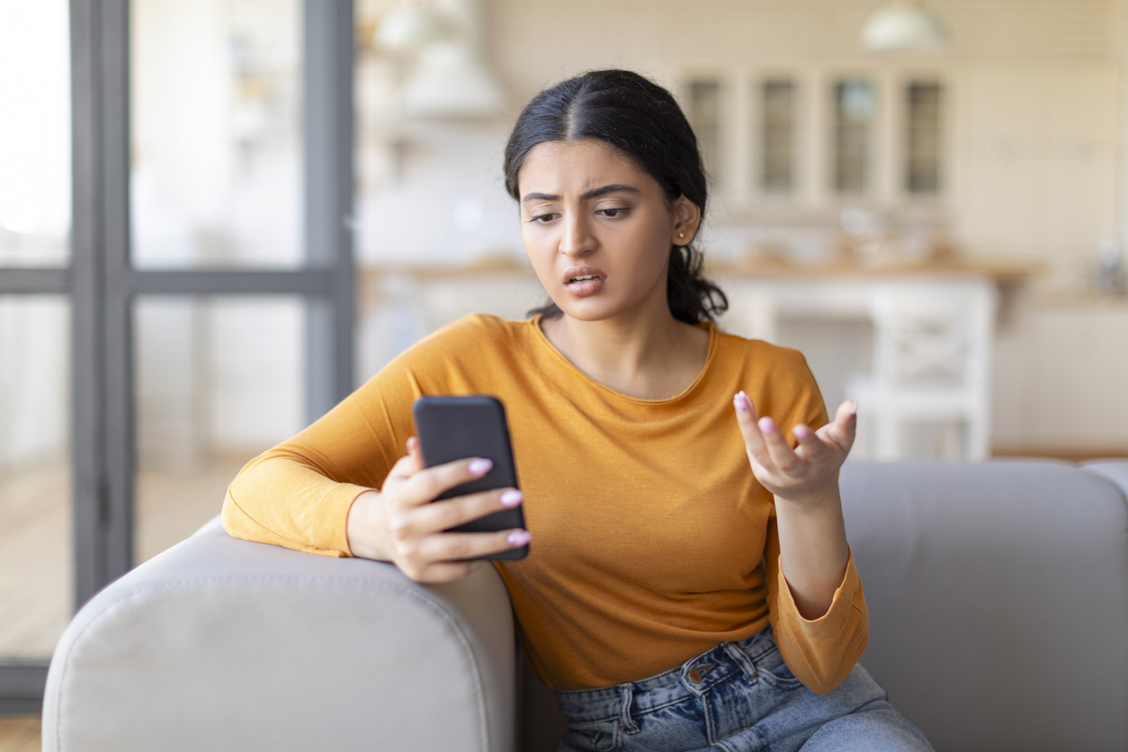 A woman sits on her couch reading a text with a frustrated look because of too much mass texting. 