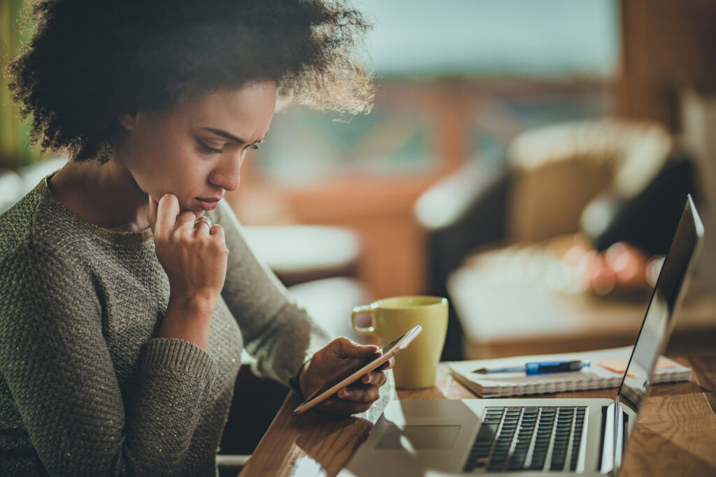 Women reads SMS message on her phone while sitting at her laptop.